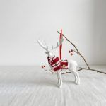 The white reindeer ornament hanging from a branch by a red ribbon, showcasing the red and white striped knit scarf against a clean white background.