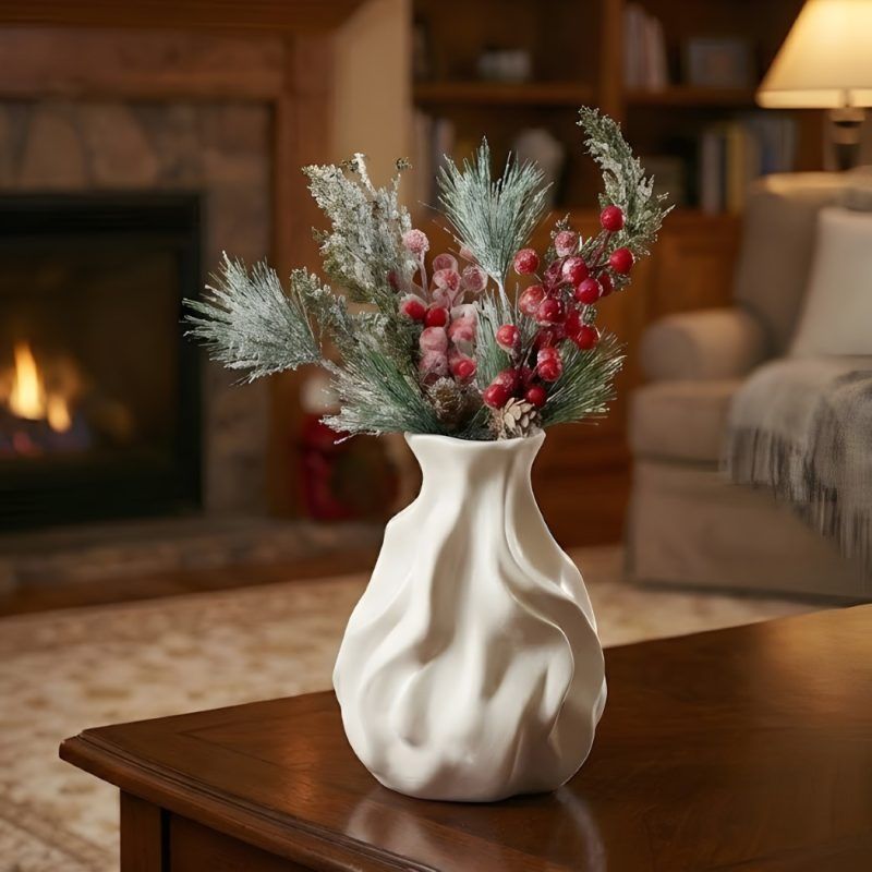 Frosted red berry and pine stems arranged in a white ceramic vase, set against a cozy, blurred fireplace background.