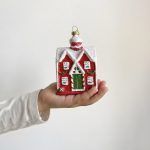 A hand holding the red cottage Christmas ornament, providing a clear reference for the size of the house ornament.