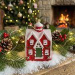 The red snowy cottage ornament placed on a decorated mantelpiece surrounded by greenery and snow, with a lit Christmas tree in the blurred background.
