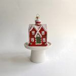 Front view of the red cottage Christmas ornament sitting on a white pedestal, showing details of the snowy roof, green door, and candy cane striped trim.