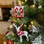 Both ornaments from the set hanging on a flocked Christmas tree; the red cottage hangs above the white reindeer with a red scarf.