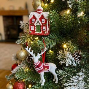 Both ornaments from the set hanging on a flocked Christmas tree; the red cottage hangs above the white reindeer with a red scarf.