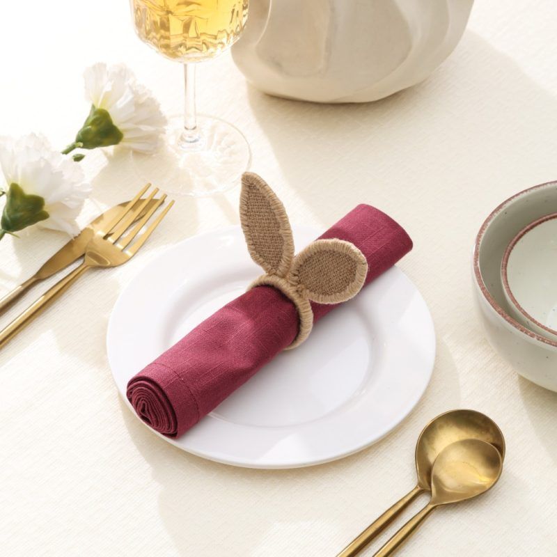 Woven rabbit ear napkin holder made of natural jute displayed on a white plate with gold cutlery.