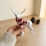 A hand holding the white reindeer ornament to show scale, highlighting the texture of the white finish and the red and white knit scarf.