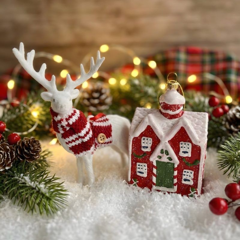 The set of two Christmas ornaments—a white reindeer and a red cottage—arranged in faux snow with pine cones and twinkling lights for a winter wonderland scene.