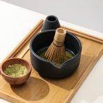 A bamboo whisk (prop) resting inside a Glimpse Homes matte black ribbed ceramic matcha bowl on a wooden tray.