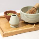 A bamboo whisk resting inside a Glimpse Homes matte mint green ribbed ceramic matcha bowl on a wooden tray.