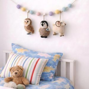 Glimpse Homes felt animal garland featuring a ram, owl, and penguin hanging above a child's bed with pastel pom-poms.