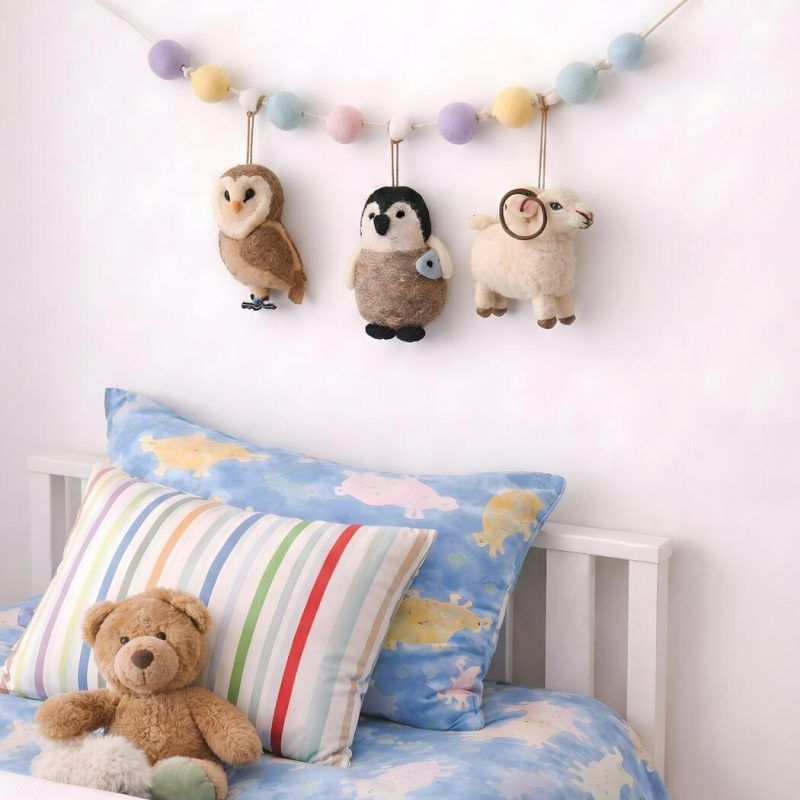 glimpse-homes-felt-animal-garland-nursery-wall-decor Glimpse Homes felt animal garland featuring a ram, owl, and penguin hanging above a child's bed with pastel pom-poms.