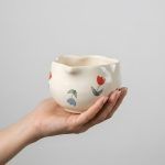 Close-up of a hand holding the Glimpse Homes floral bloom matcha bowl, highlighting the red and blue tulip pattern and pouring spout.