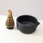 Angled closeup showing the pouring spout and ribbed design of the Glimpse Homes matte black matcha bowl next to a whisk on its ceramic holder.