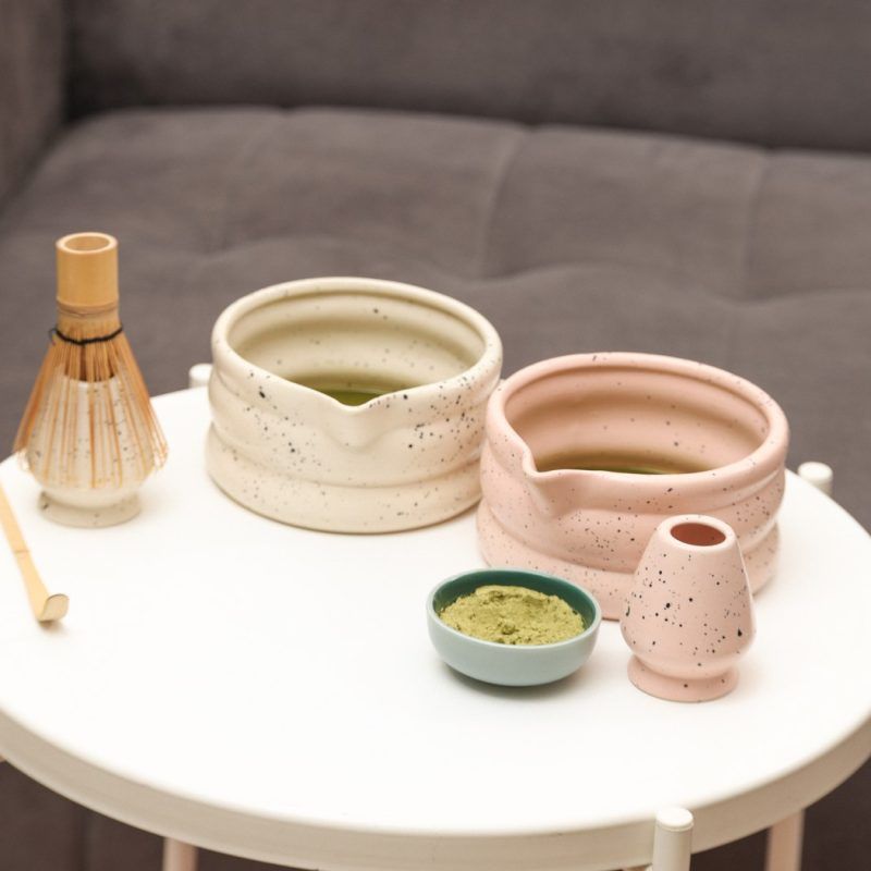 Glimpse Homes matte pink and ivory speckled ceramic matcha bowls filled with green tea, displayed with matching whisk holders and accessories on a white table.