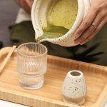 Action shot of pouring green tea from a Glimpse Homes matte off-white speckled matcha bowl into a glass cup, highlighting the pouring spout.