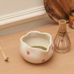 Freshly prepared green tea served in a Glimpse Homes white ceramic bowl with red floral patterns and a scalloped rim, next to a whisk holder.