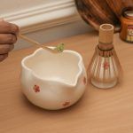 Hand adding green matcha powder to a Glimpse Homes red floral ceramic bowl using a bamboo scoop, with the matching whisk holder nearby.