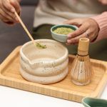 Person adding matcha powder with a bamboo scoop into a Glimpse Homes matte off-white speckled ceramic bowl with matching whisk holder nearby.