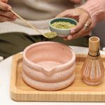 Person adding green matcha powder to a Glimpse Homes matte pink speckled ceramic bowl using a bamboo scoop, with the whisk holder nearby.