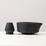 Side profile view of a Glimpse Homes matte black matcha bowl with horizontal ribbed texture and a matching ceramic whisk stand.
