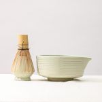 Side profile view of a Glimpse Homes matte mint green matcha bowl with horizontal ribbed texture and a matching ceramic whisk stand.