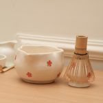 Side profile of the Glimpse Homes white ceramic matcha bowl featuring red floral patterns, shown next to a bamboo whisk on a stand (whisk is a prop).
