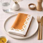 Close-up of Glimpse Homes Terracotta Bloom napkin showing the printed white lace-style border on 100% cotton fabric.