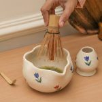 Hand using a bamboo whisk to froth green tea inside a Glimpse Homes floral bloom ceramic matcha bowl with a scalloped rim.