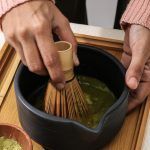 Hands using a bamboo whisk to froth green tea inside a Glimpse Homes matte black ribbed ceramic matcha bowl with a pouring spout.