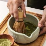 Hands using a bamboo whisk to froth green tea inside a Glimpse Homes matte mint green ribbed ceramic matcha bowl with a pouring spout.