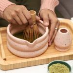 Hands using a bamboo whisk to froth green tea inside a Glimpse Homes matte pink speckled matcha bowl with a unique puffy rim design.