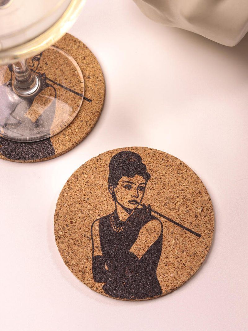Close up of a Glimpse Homes 4-inch round cork coaster featuring a vintage Audrey Hepburn illustration, sitting next to a wine glass on a bright surface.