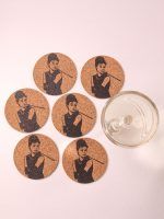A complete set of six Glimpse Homes 4-inch cork coasters with an Audrey Hepburn illustration, arranged in a circular pattern next to an empty glass.