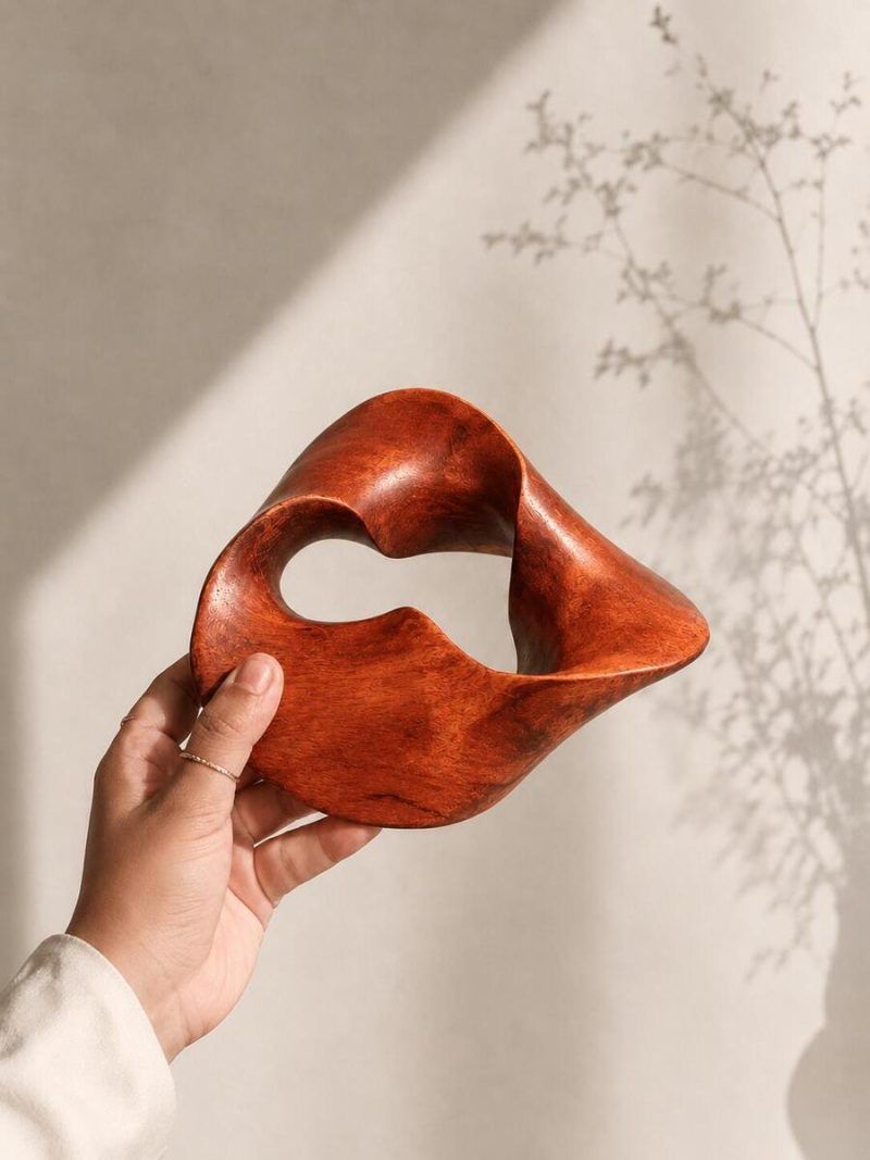 A hand holding the handcrafted wooden Trinity Knot centrepiece against a wall with delicate plant shadows, highlighting the smooth matte mahogany finish and organic wood grain.