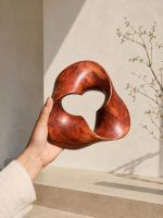 A hand holding The Trinity Knot abstract wooden shelf decor, showcasing its lightweight design and 7.5-inch size against a warm beige background with botanical shadows.