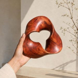 A hand holding The Trinity Knot abstract wooden shelf decor, showcasing its lightweight design and 7.5-inch size against a warm beige background with botanical shadows.