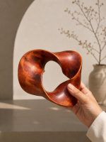 Close-up of a hand holding The Trinity Knot, an endless loop wooden showpiece by Glimpse Homes, with an arched wall and ceramic vase in the background.