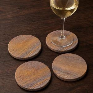 Four Glimpse Homes natural sandstone coasters arranged on a dark wooden table, with one coaster elegantly holding a glass of white wine to protect the surface.