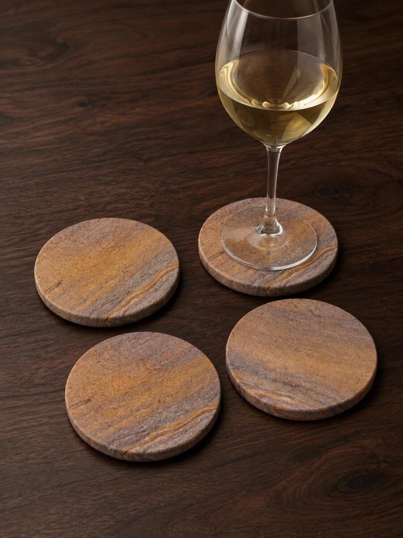 Four Glimpse Homes natural sandstone coasters arranged on a dark wooden table, with one coaster elegantly holding a glass of white wine to protect the surface.