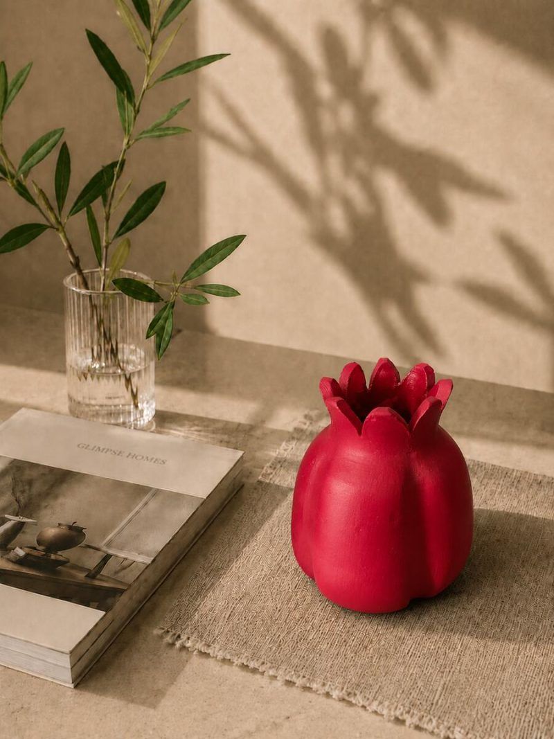 Large handmade terracotta pomegranate showpiece in a ravishing red finish, set on a woven mat as aesthetic table decor alongside a Glimpse Homes book.