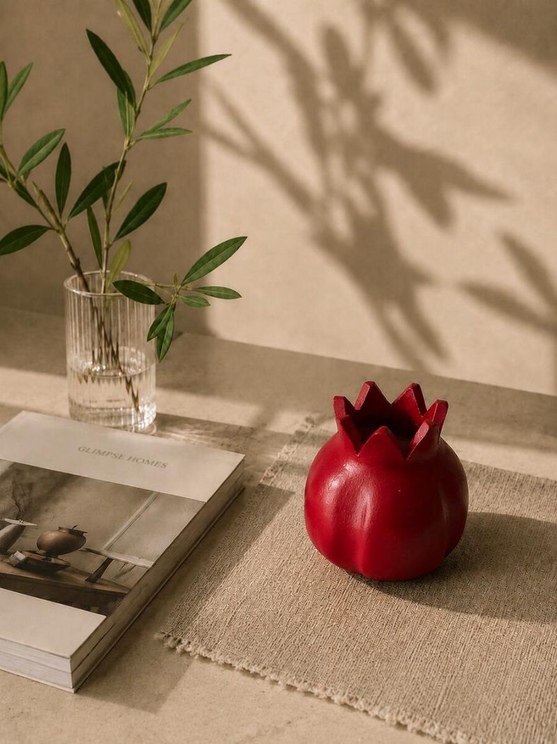 Handmade terracotta pomegranate table decor in ravishing red, placed on a woven mat next to a Glimpse Homes book and an olive branch vase.