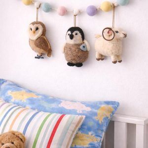 Glimpse Homes felt animal garland featuring a ram, owl, and penguin hanging above a child's bed with pastel pom-poms.