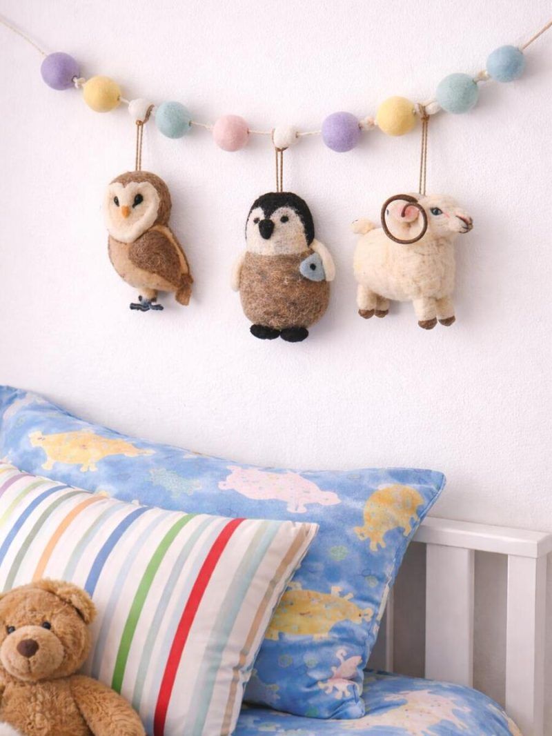 Glimpse Homes felt animal garland featuring a ram, owl, and penguin hanging above a child's bed with pastel pom-poms.